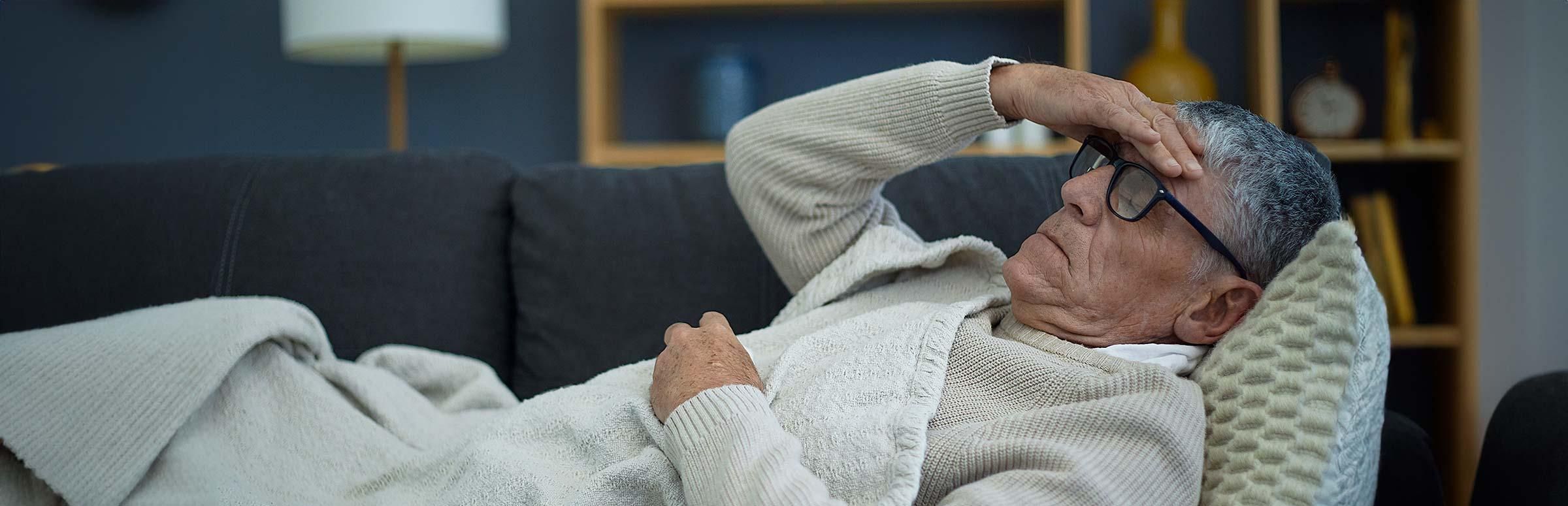 Grippe: Ein kranker Mann liegt auf dem Sofa und greift sich an den Kopf.