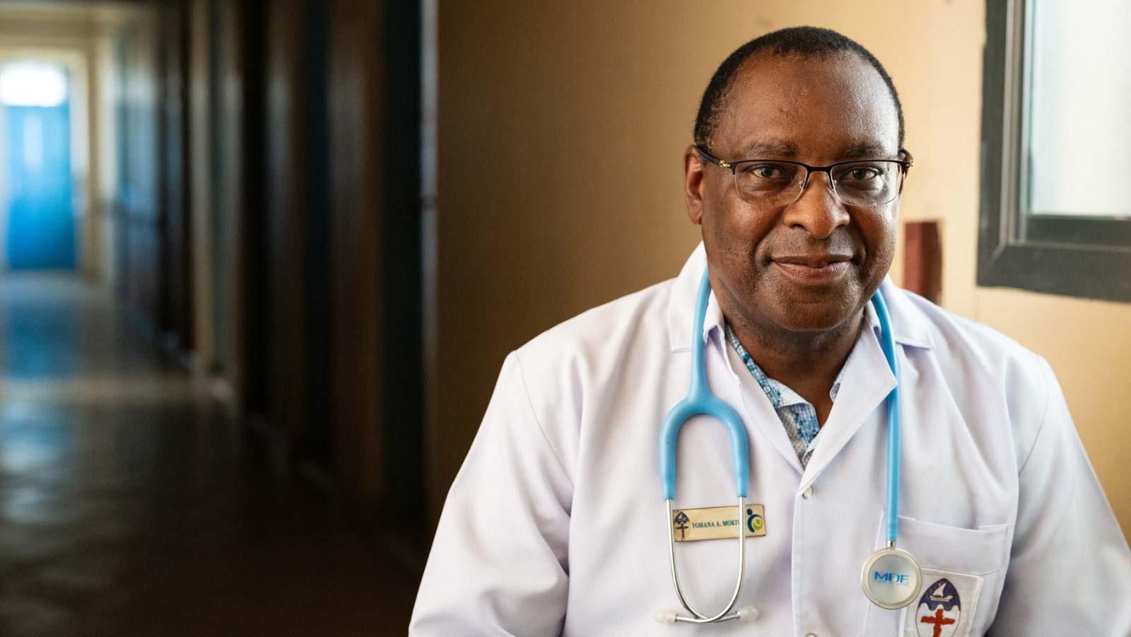 Dr. Yohana Mokiwa, ein lächelnder Mann mittleren Alters mit kurzen Haaren und Brille, in Arztkleidung. 